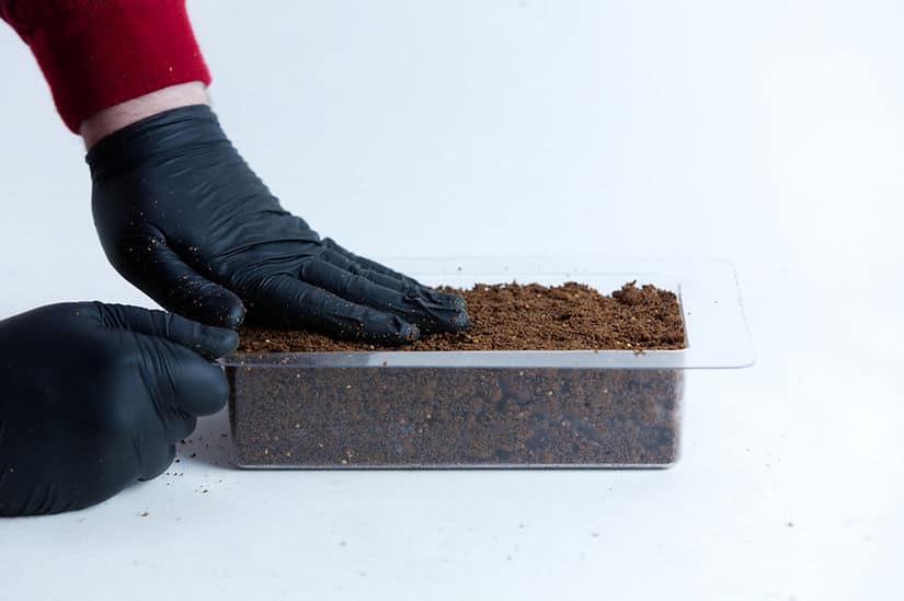 Mycelium Mold Growth form -Brick (Reusable) 6 Hands in black gloves press soil into the Mycelium Mould Growth form - Brick (Reusable), shown in a clear plastic container against a white background.