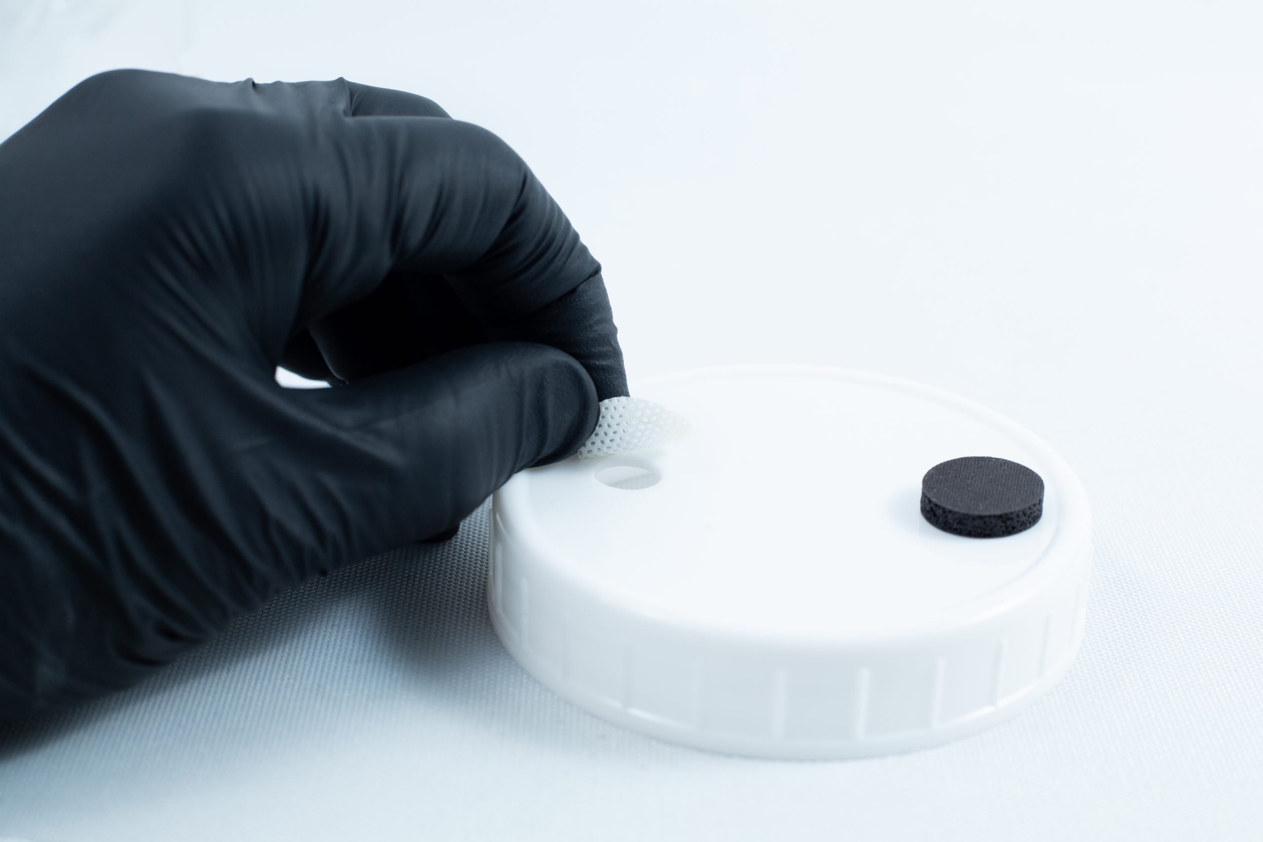 Primer 94 multisurface for 3M glue 10ML 5 A gloved hand positions a mesh disc into a hole on a white lid beside a black disc, preparing the area for adhesion using Primer 94 multisurface for 3M glue 10ml.