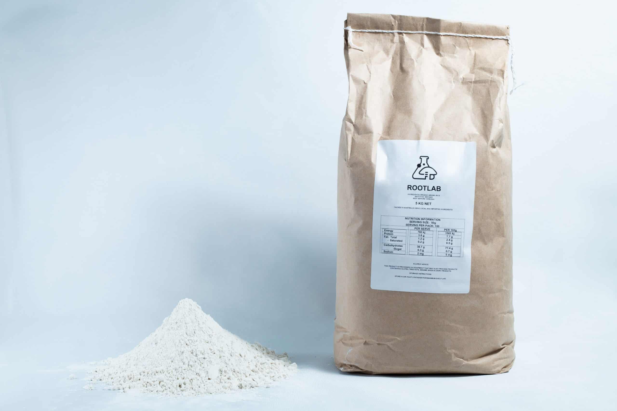 Brown Rice Flour 5kg Organic Food Grade 1 A bag labelled ROOTLAB, containing Brown Rice Flour 5kg Organic Food Grade, stands beside a mound of fine white powder on a white background.