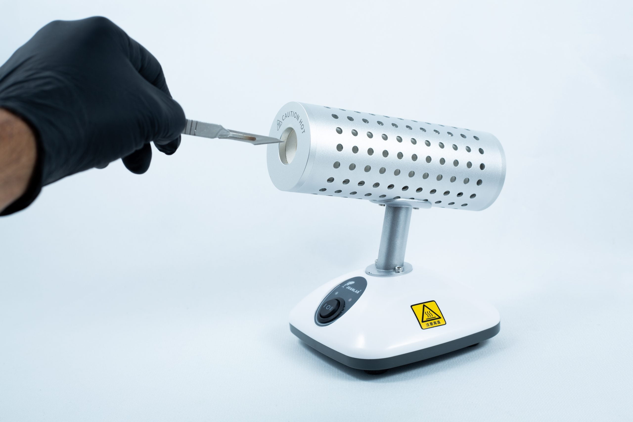 Infrared Sterilizer Bacti Cinerator Lab tool 4 A gloved hand inserts a tool into the Infrared Steriliser Bacti Cinerator Lab tool, which features a warning sign on its base, in the laboratory.