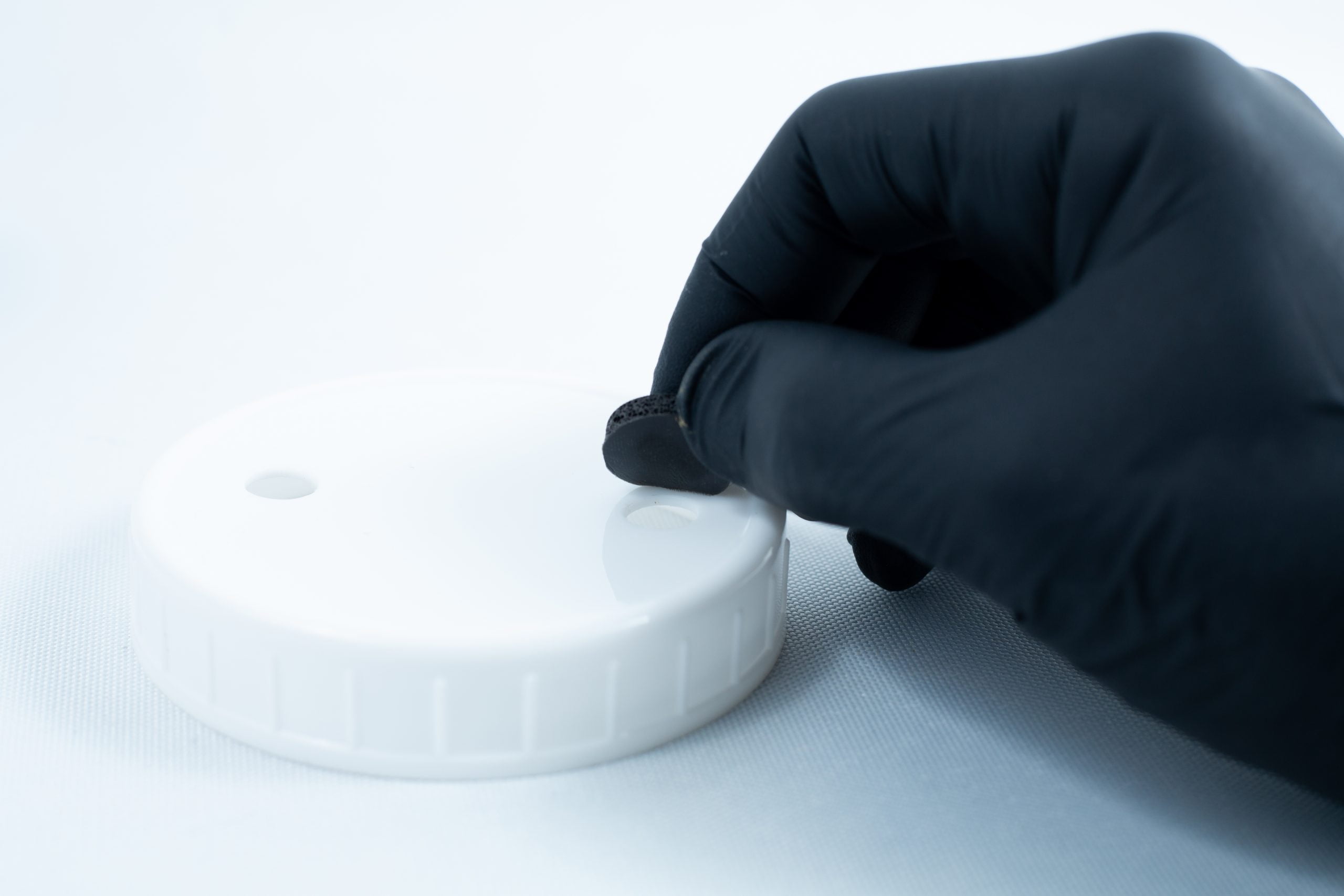Primer 94 multisurface for 3M glue 10ML 4 A gloved hand applies Primer 94 multisurface for 3M glue 10ML onto a round white plastic lid to prepare it for optimal tape adhesion.