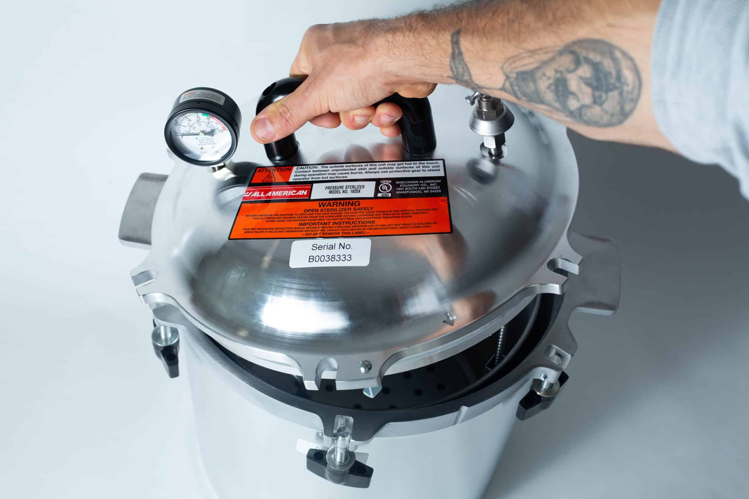 1925x All-American Sterilizer (Autoclave) 25 quart/ 24 Liter 2 A tattooed hand holding the lid of a stainless steel pressure cooker with a gauge and warning labels.