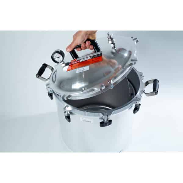 A hand lifts the lid of a large silver pressure cooker with a gauge and black handles.