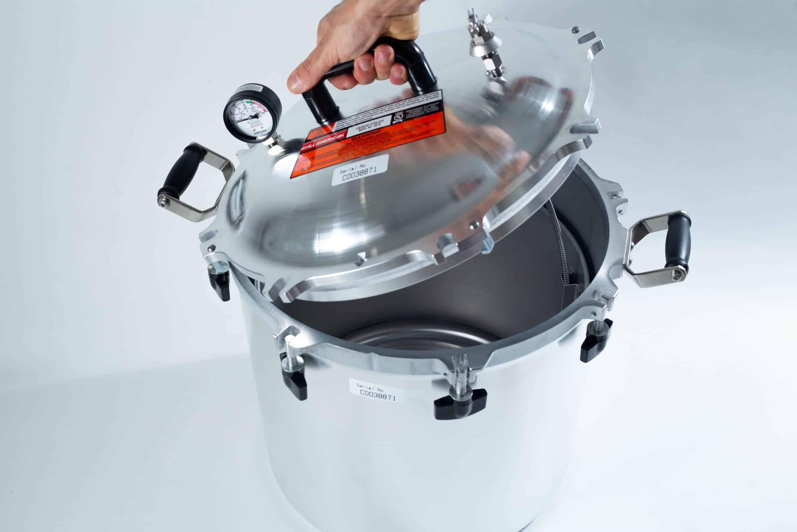 1941x All-American Sterilizer (Autoclave) 41 quart/ 39 liters. 2 A hand lifts the lid of a large silver pressure cooker with a gauge and black handles.