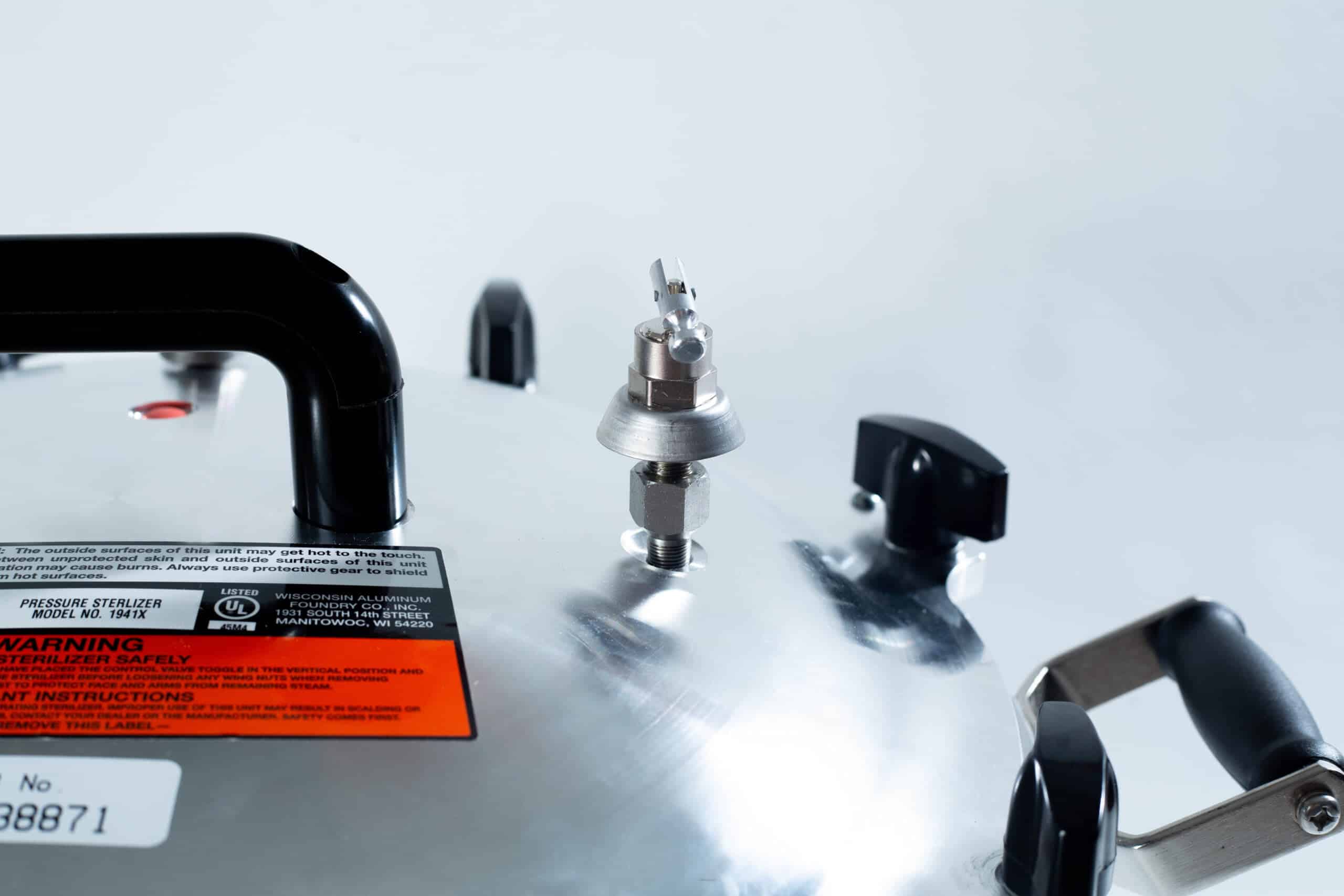 1941x All-American Sterilizer (Autoclave) 41 quart/ 39 liters. 7 Close-up of a metallic pressure cooker lid with handles, safety valve, and warning label visible.
