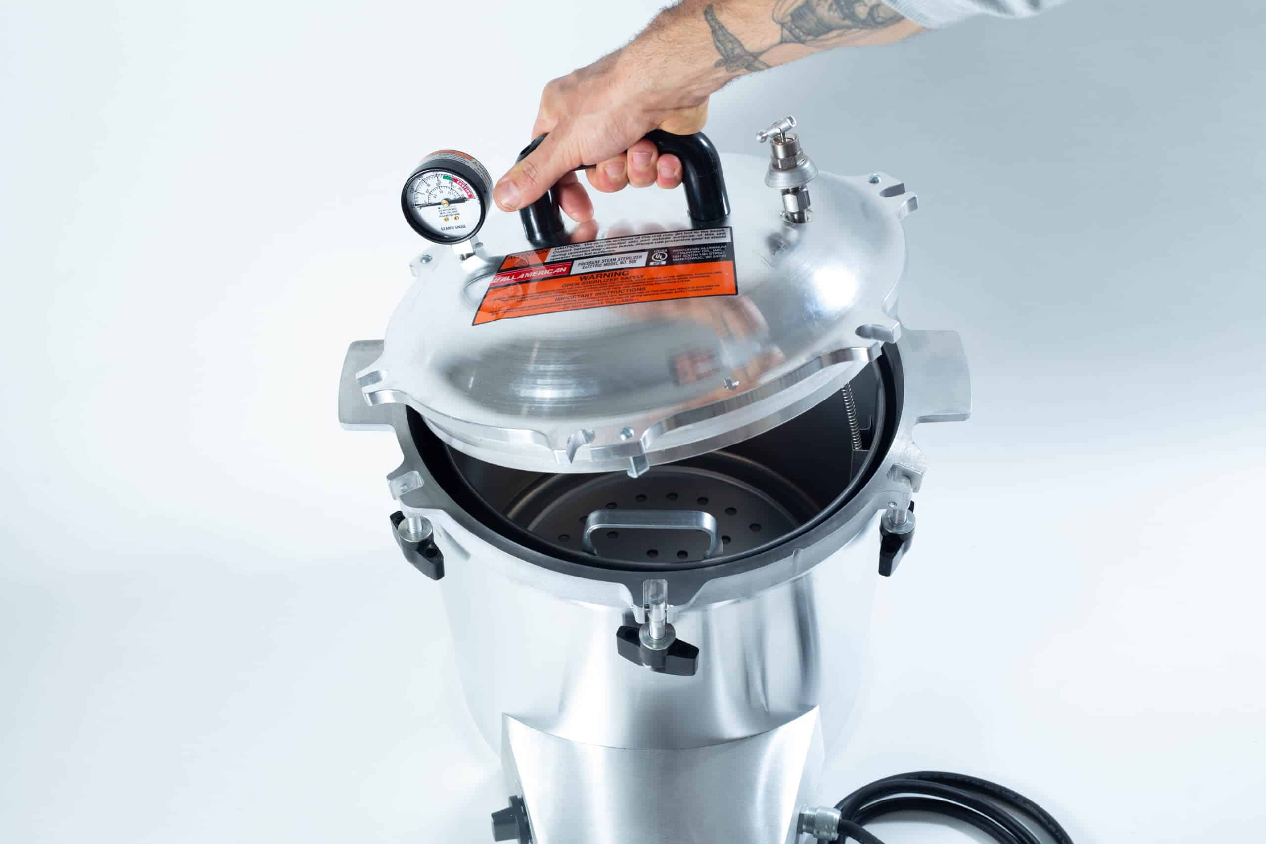 50x All American electric autoclave sterilizer 25 quart 3 A hand lifts the lid of a stainless steel pressure cooker with a gauge and safety label on top.
