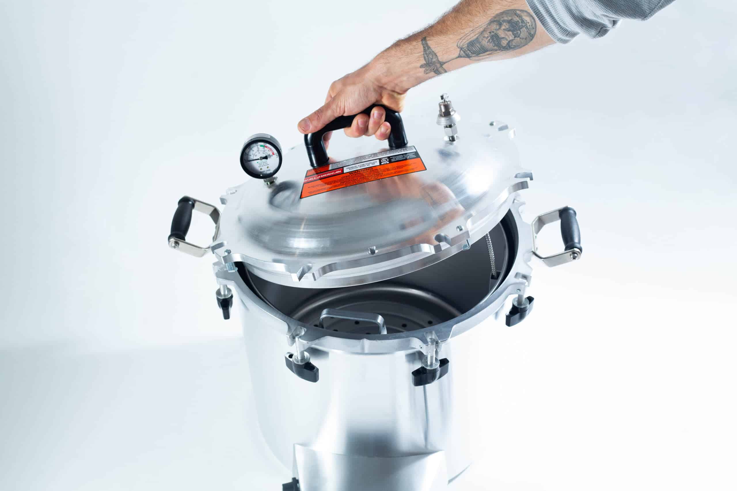75x All American electric autoclave sterilizer 41 quart 3 A hand lifts the lid of a stainless steel pressure cooker with a gauge on top.