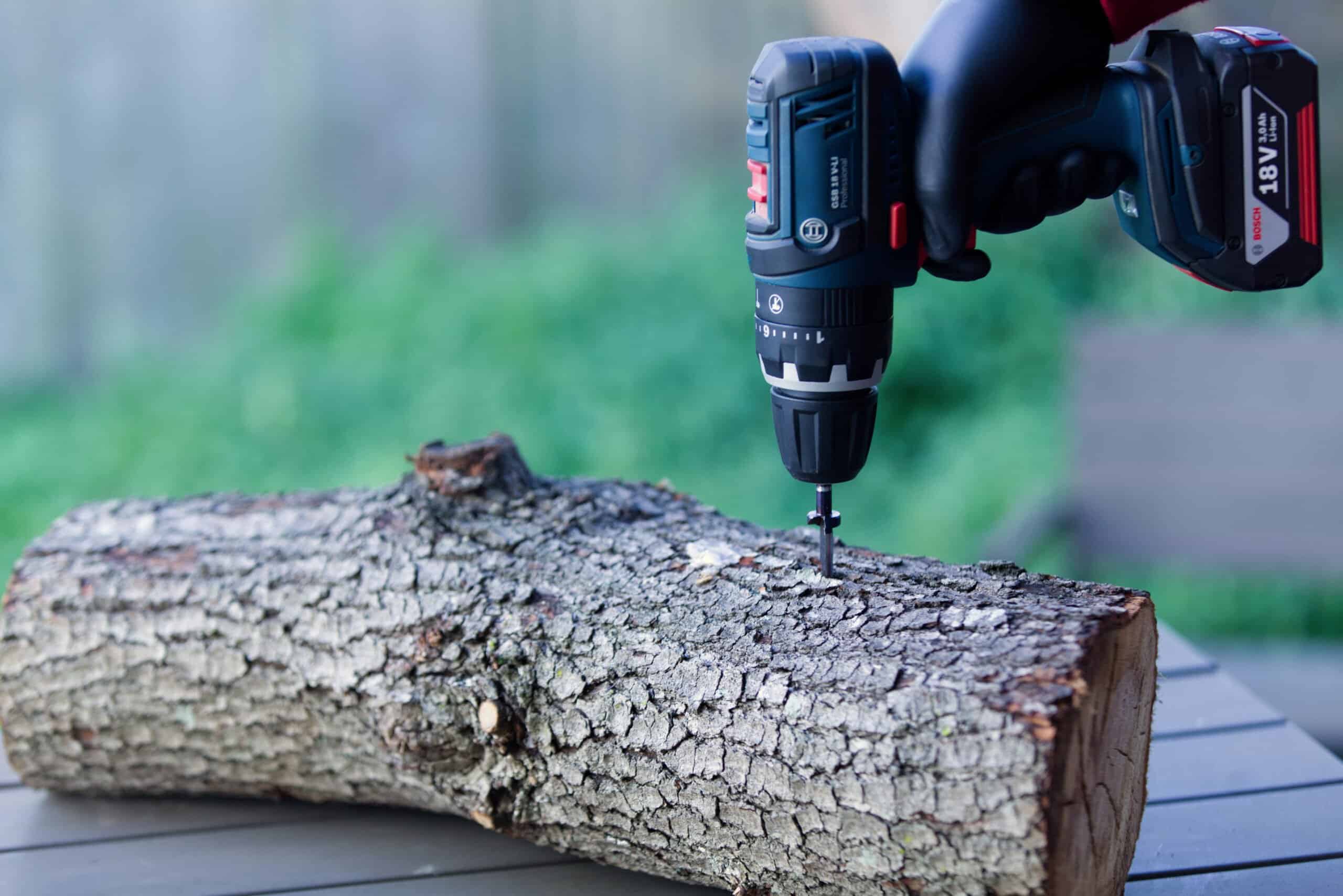 Mushroom Log Kit | Mushroom Inoculation Kit (Log Dowel) 3 A gloved hand uses a power drill to drive a screw into a log on an outdoor table.