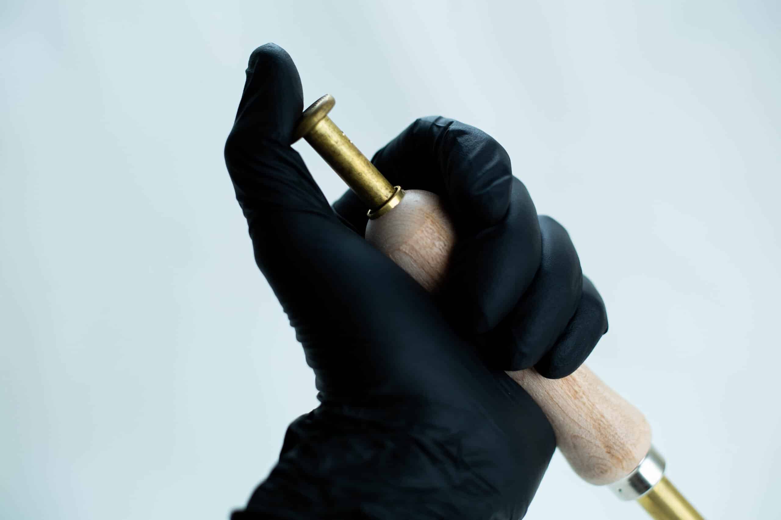 Nitrile Powder Free Disposable Gloves 3 A hand in a black glove holding a brass and wooden tool against a plain light background.