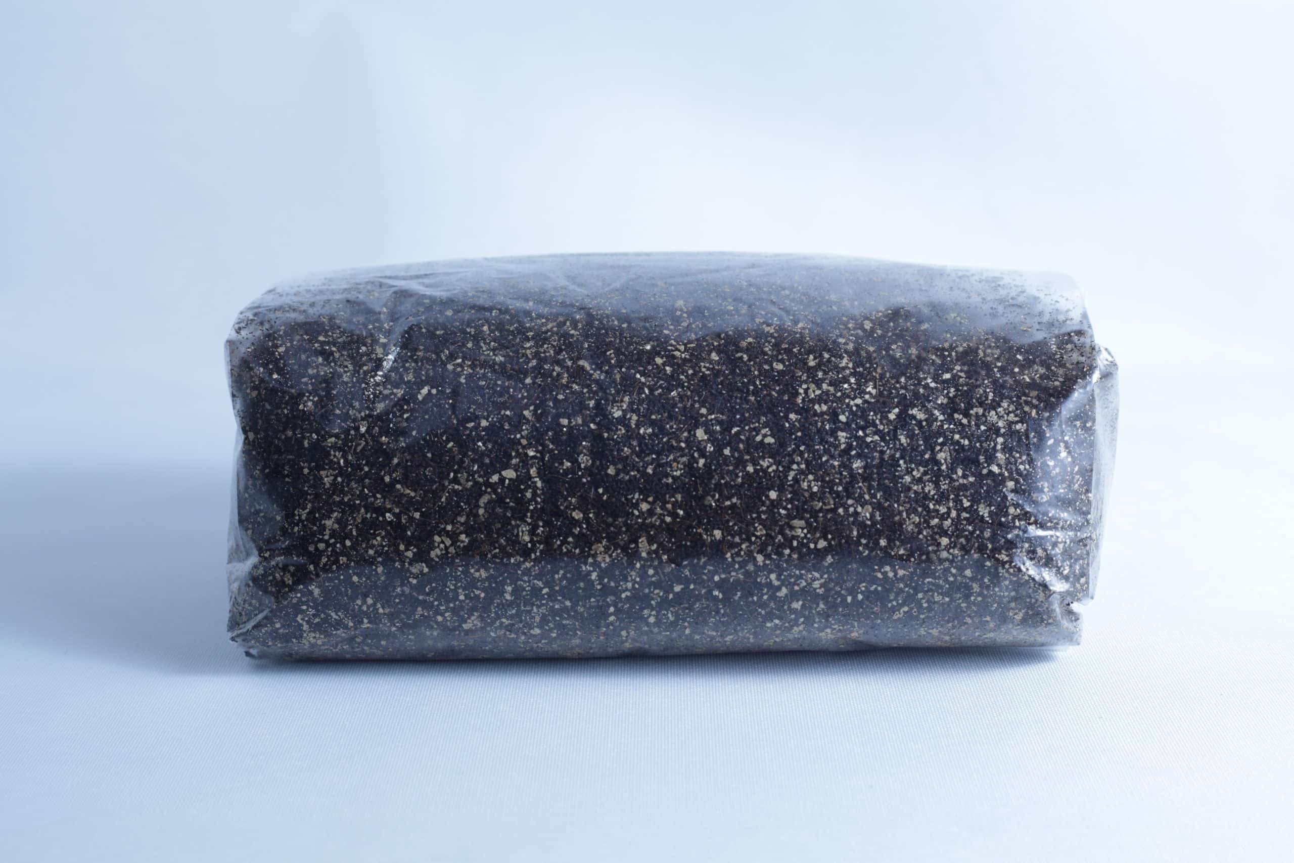 Monotub Mushroom Growing Kit-Premium 9 A block of dark soil or compost, perfect for the Monotub Mushroom Growing Kit-Premium, is wrapped in clear plastic and set against a plain light background.