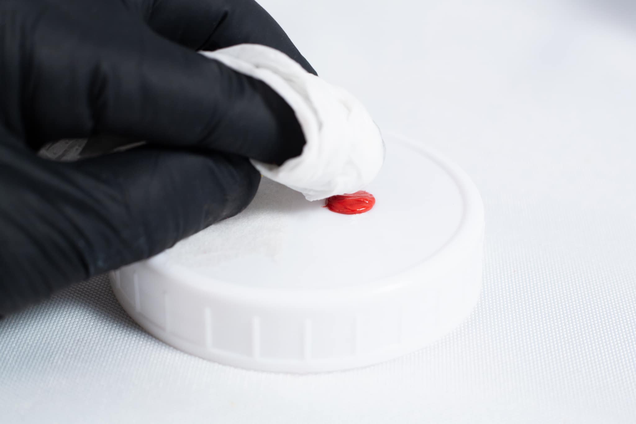 High Temperature RTV Silicon-Red 6 A gloved hand wipes a red stain from a white plastic lid with a tissue, ensuring the surface is clean before applying High Temperature RTV Silicone-Red.
