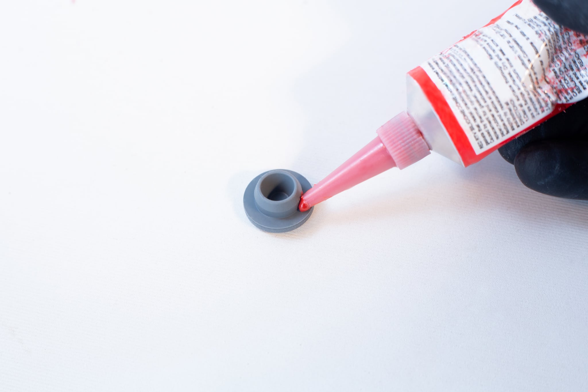 High Temperature RTV Silicon-Red 2 A hand applies High Temperature RTV Silicone-Red from a tube onto a small grey plastic part placed on a white surface.