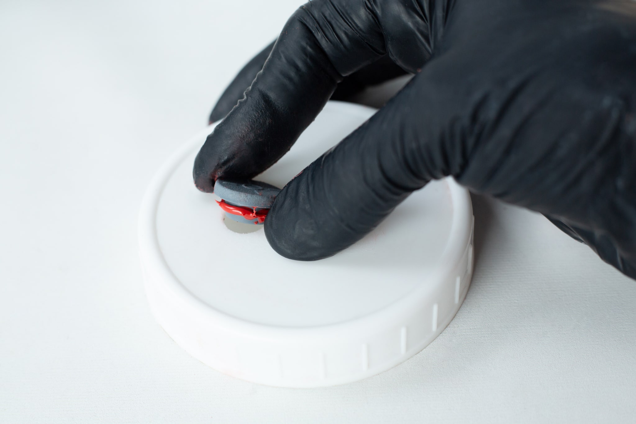 High Temperature RTV Silicon-Red 3 A gloved hand holds a small round object above a white plastic lid, ready to apply High Temperature RTV Silicone-Red on a light surface.