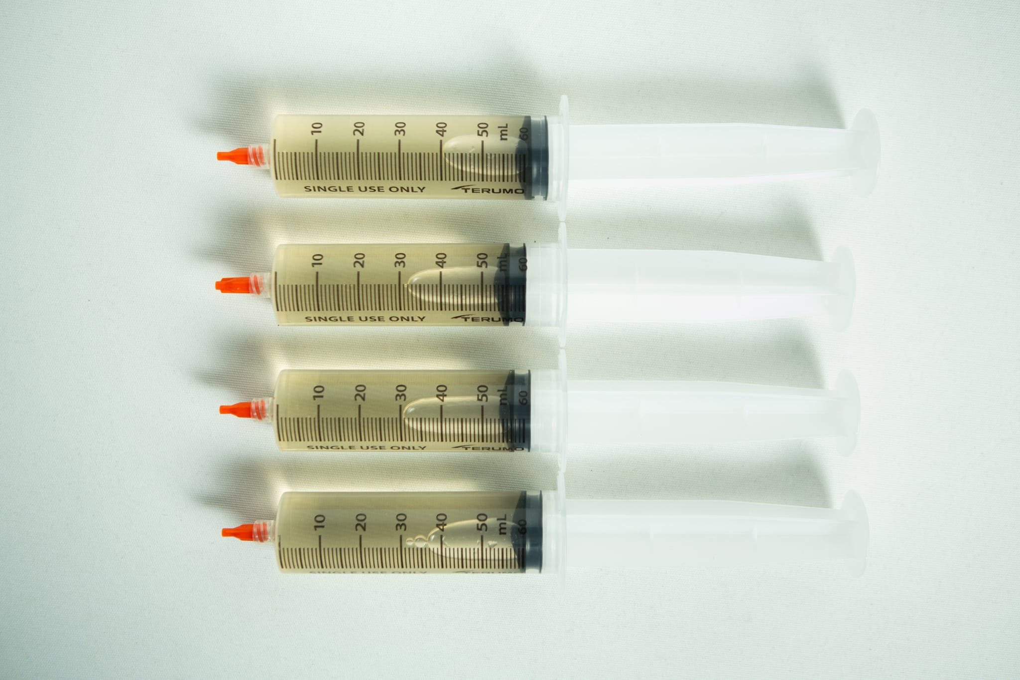 Liquid culture to Liquid culture expansion Kit 3 Four large syringes with orange caps from the Liquid culture to Liquid culture expansion Kit are lined up horizontally on a white surface.
