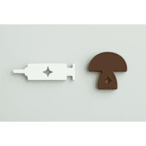 A white Syringe Opener and a brown mushroom-shaped piece, each with diamond-shaped cut-outs, rest on a light background.