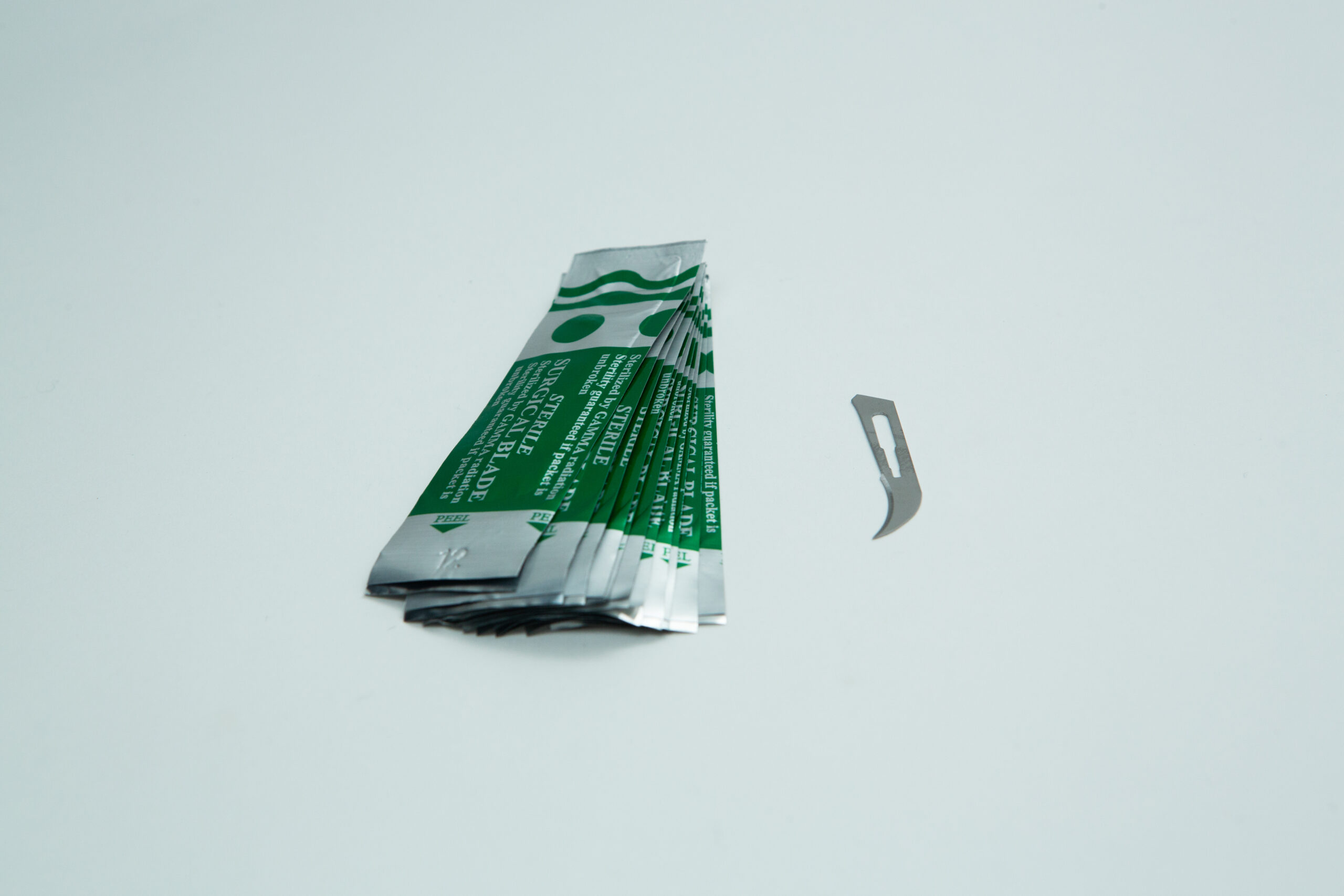 Scalpel handle #3 and blades | Mycology 5 Five green and silver foil packets lie next to a single small, curved metal blade on a white surface.