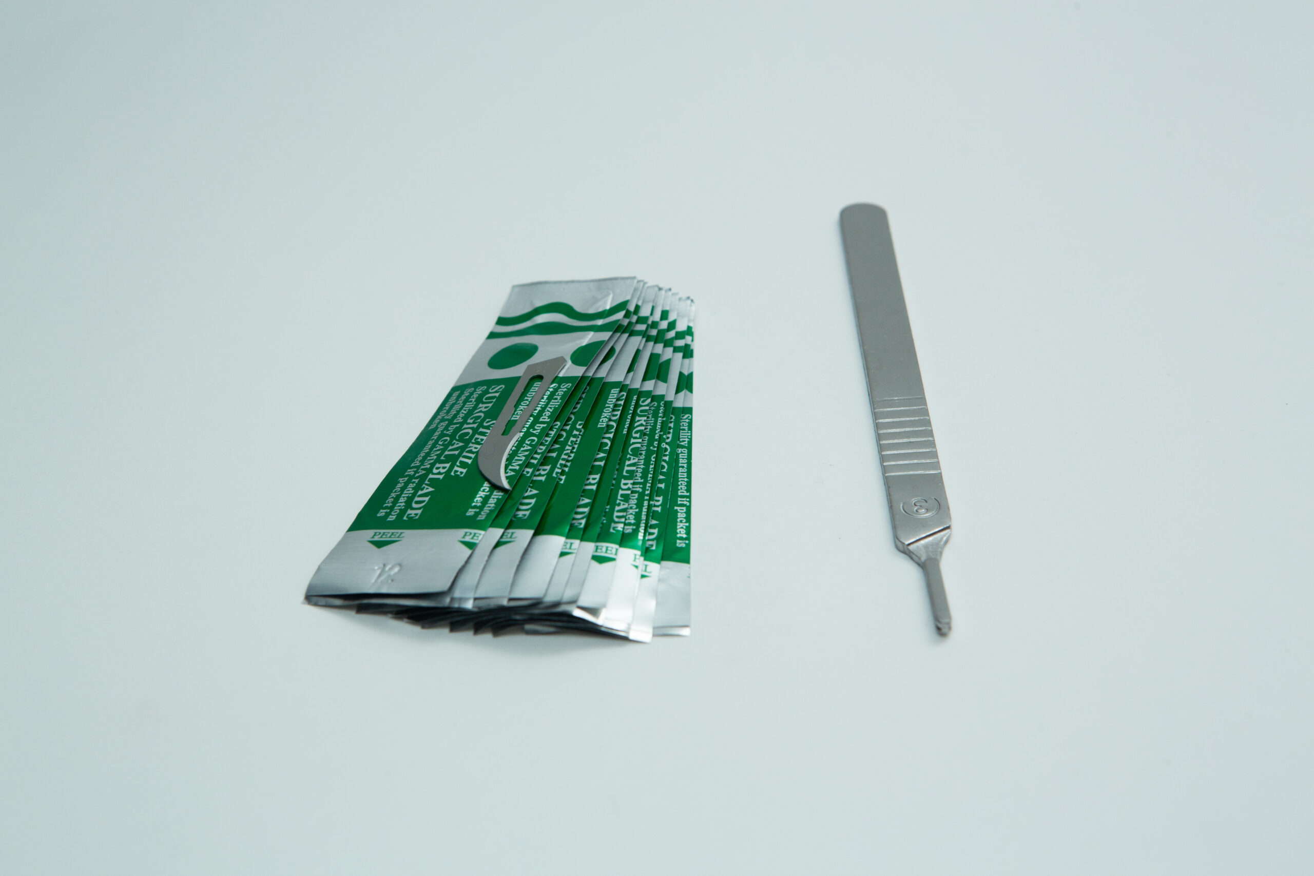 Scalpel handle #3 and blades | Mycology 6 A metal scalpel handle and several individually wrapped scalpel blades on a white background.