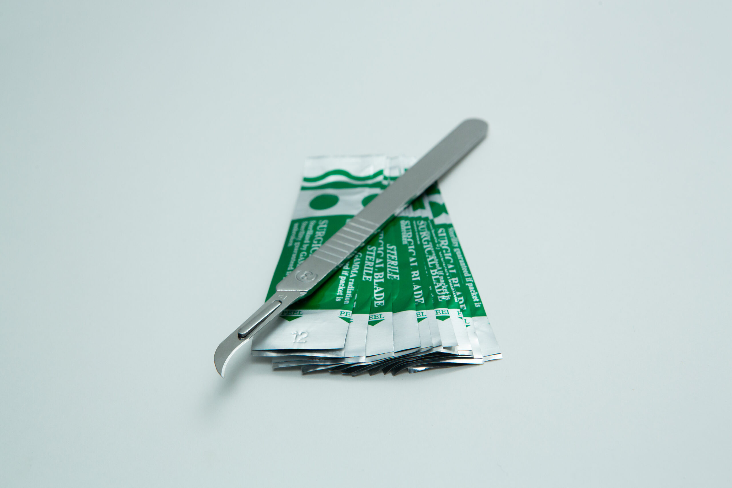Scalpel handle #3 and blades | Mycology 7 A scalpel rests on several green and silver sterile blade packets on a white background.