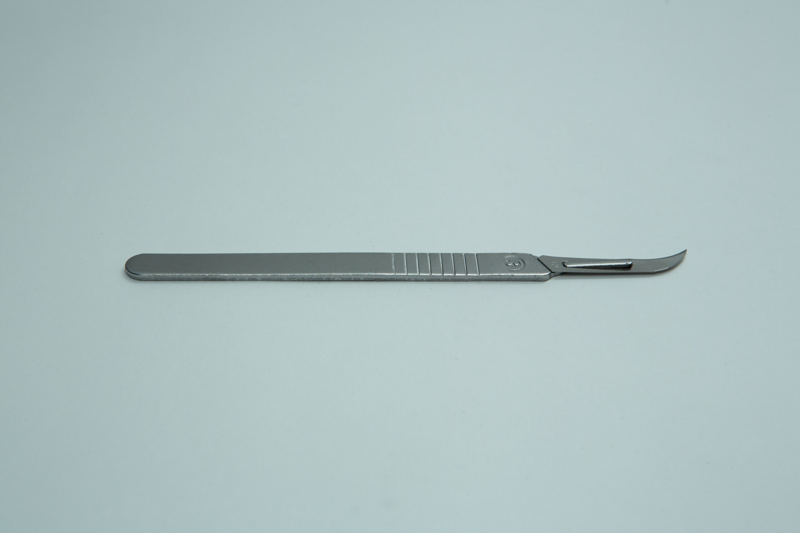 Scalpel handle #3 and blades | Mycology 8 A metal surgical scalpel with a curved blade lies on a plain light grey background.