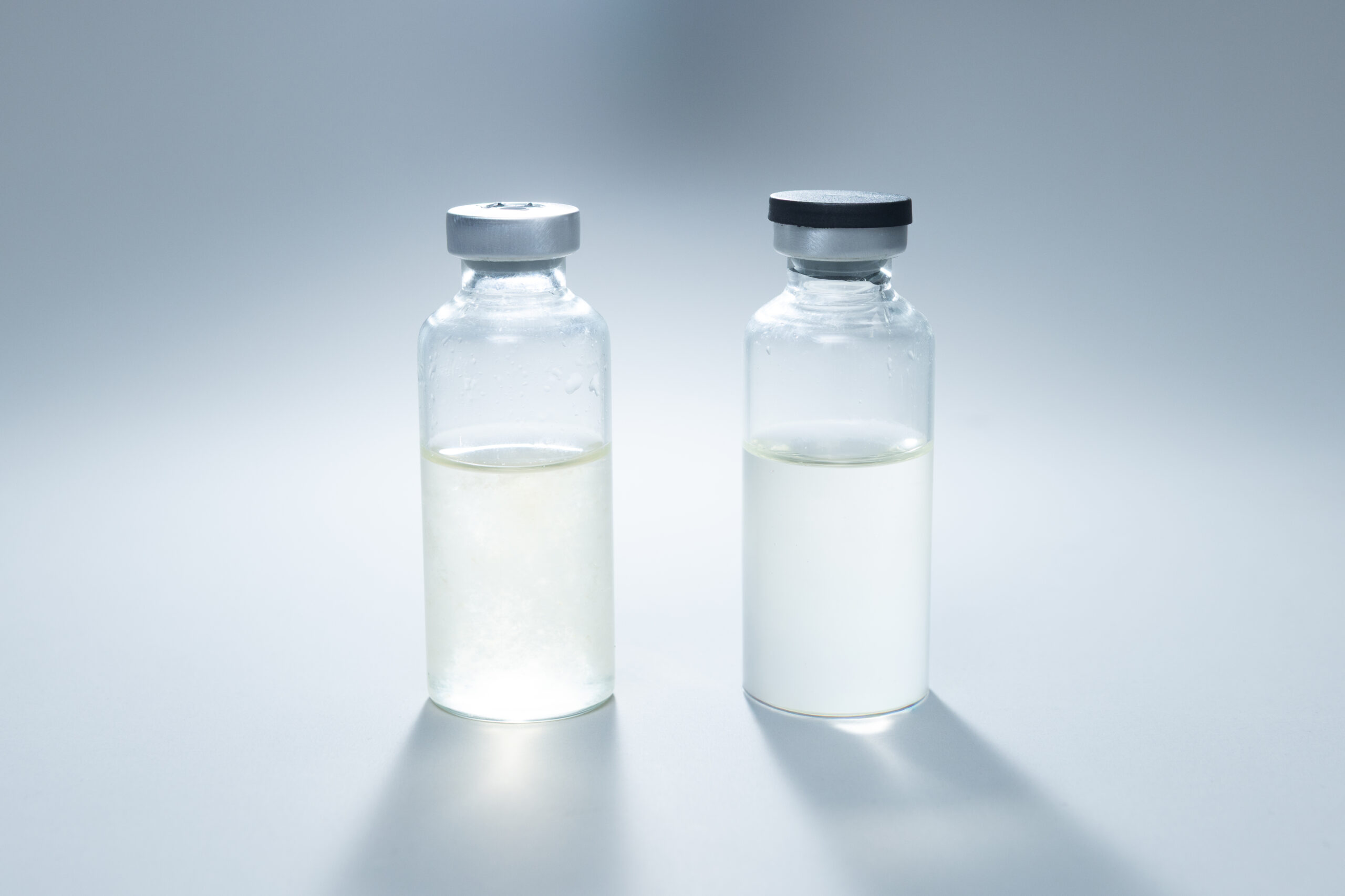 Sterile Liquid Culture Vials, Dr Myc Adaptive LC, Crimp Sealed 4 Two clear glass vials containing differently coloured liquids rest on a white surface, softly illuminated. These Dr Myc Adaptive LC Sterile Liquid Culture Vials (30 ml, crimp sealed) require no pressure cooker and are designed to ensure purity and prevent contamination.